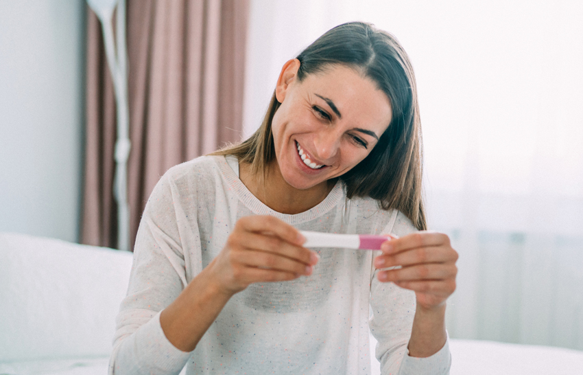 Cheerful young woman sitting on the bed and looking at pregnancy test. Happy young woman holding in hands positive pregnancy test in bedroom.