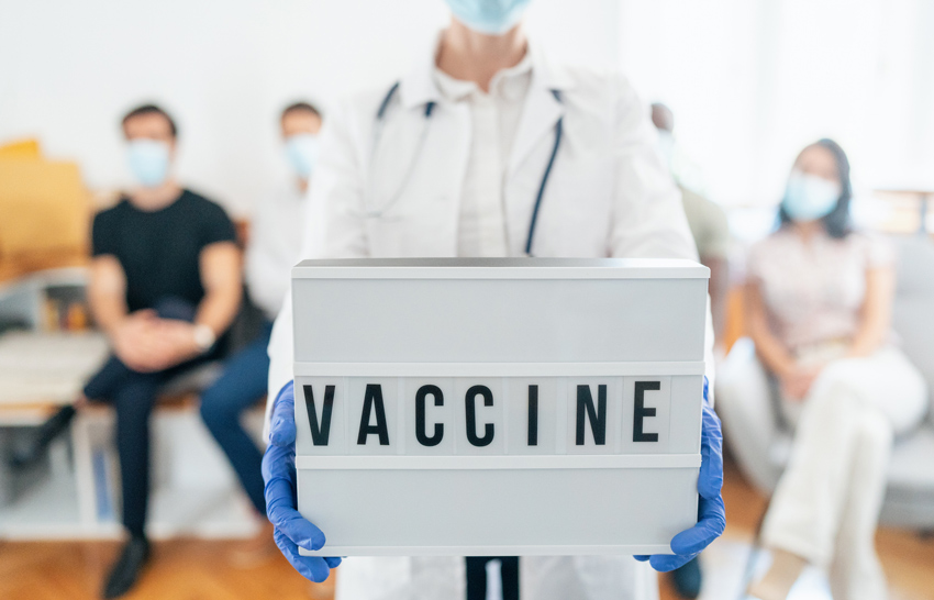 Healthcare worker with face mask showing placard with "Vaccine", people waiting to be vaccinated at vaccinating center.