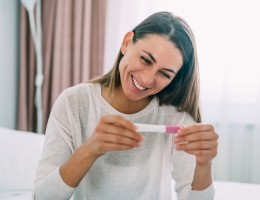 Cheerful young woman sitting on the bed and looking at pregnancy test. Happy young woman holding in hands positive pregnancy test in bedroom.