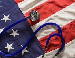 A stethoscope medical instrument resting on American flag fabric of red, white, and blue stars and stripes, representing concepts of healthcare and medicine reform, USA political changes affecting medical costs and health insurance benefit social issues, and ideas of listening, checking for healthy vital signs.
