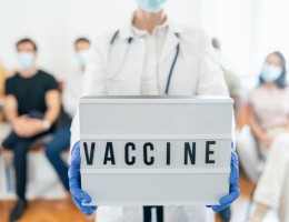 Healthcare worker with face mask showing placard with "Vaccine", people waiting to be vaccinated at vaccinating center.
